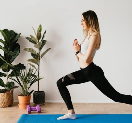 Side view of fit lady in sports clothes and wristwatch standing in Anjaneyasana pose while practicing yoga on mat near dumbbells and potted plants at home and looking away