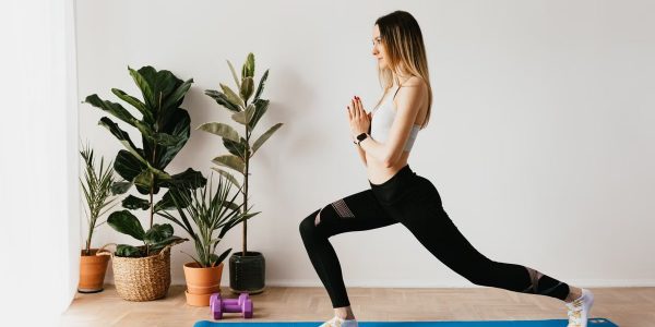 Side view of fit lady in sports clothes and wristwatch standing in Anjaneyasana pose while practicing yoga on mat near dumbbells and potted plants at home and looking away