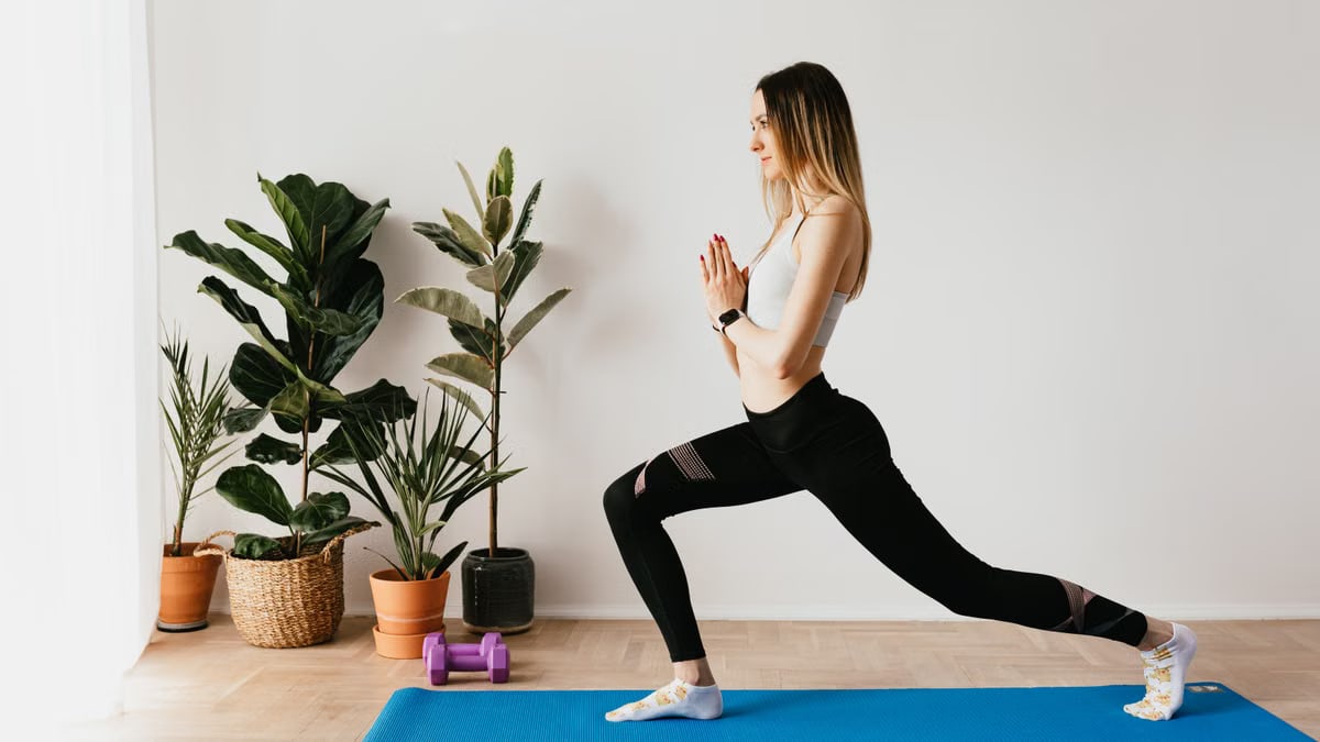 Side view of fit lady in sports clothes and wristwatch standing in Anjaneyasana pose while practicing yoga on mat near dumbbells and potted plants at home and looking away